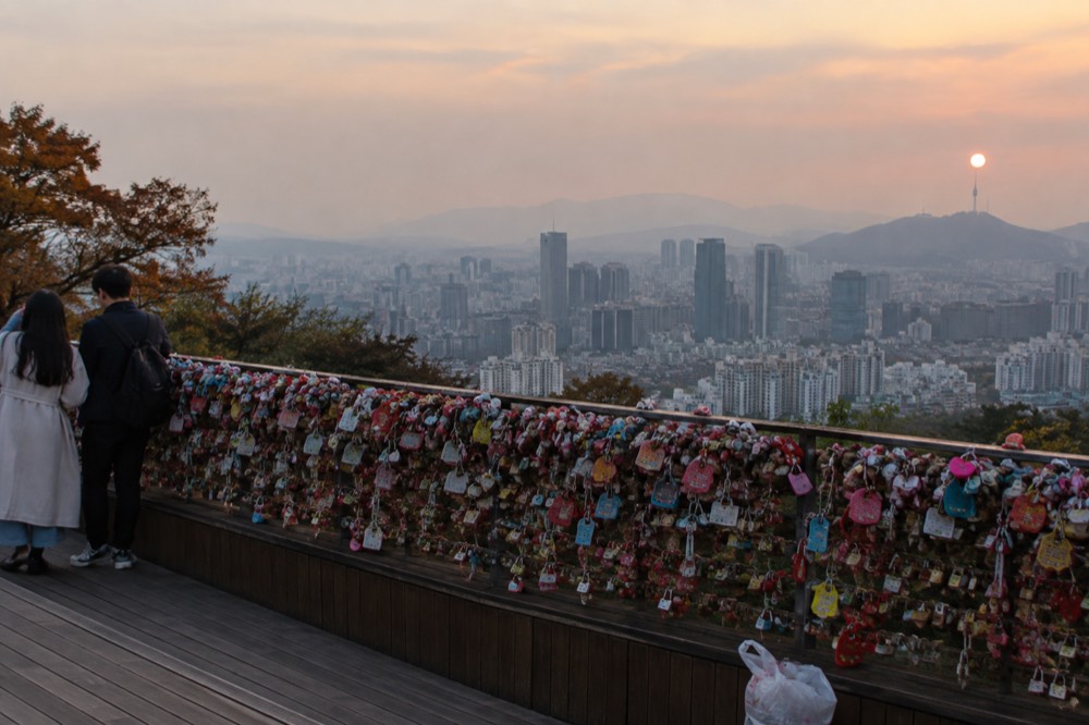 A terrace railing covered in padlocks with the Seoul skyline visible beyond