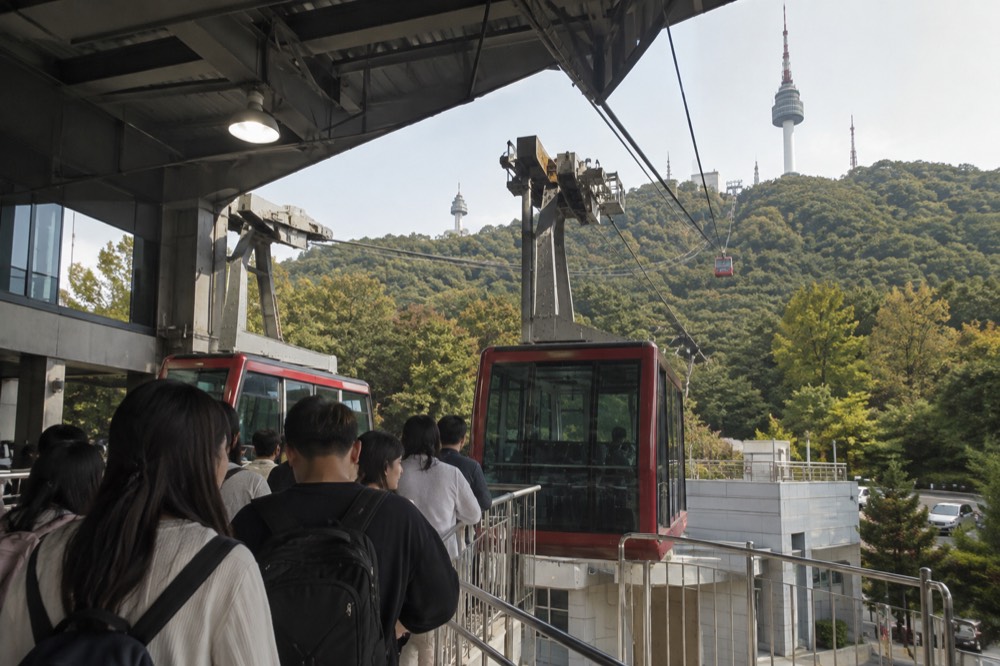 A queue of visitors waiting at the Namsan cable car platform with the tower visible above the trees