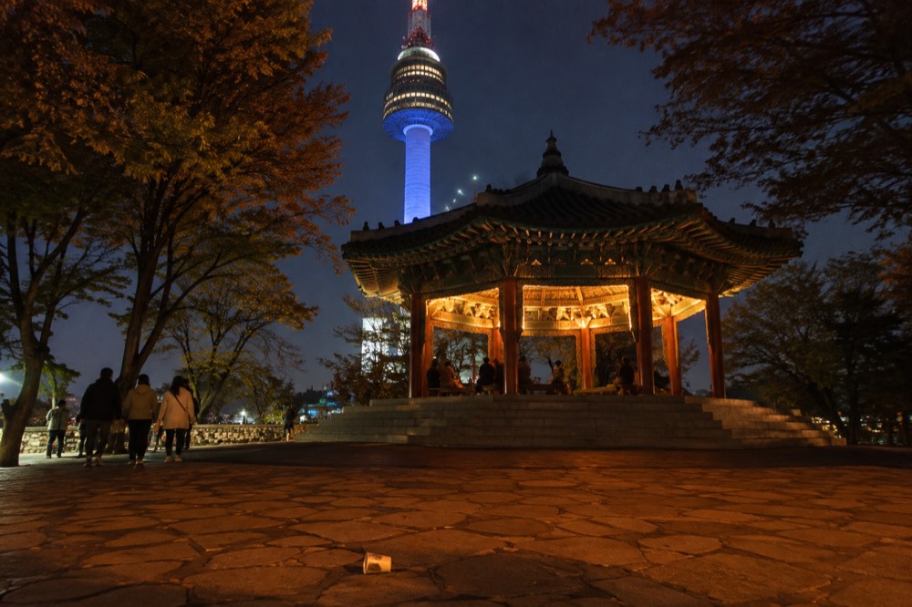 The illuminated tower rising above the lit summit plaza and the dark ridge of Namsan