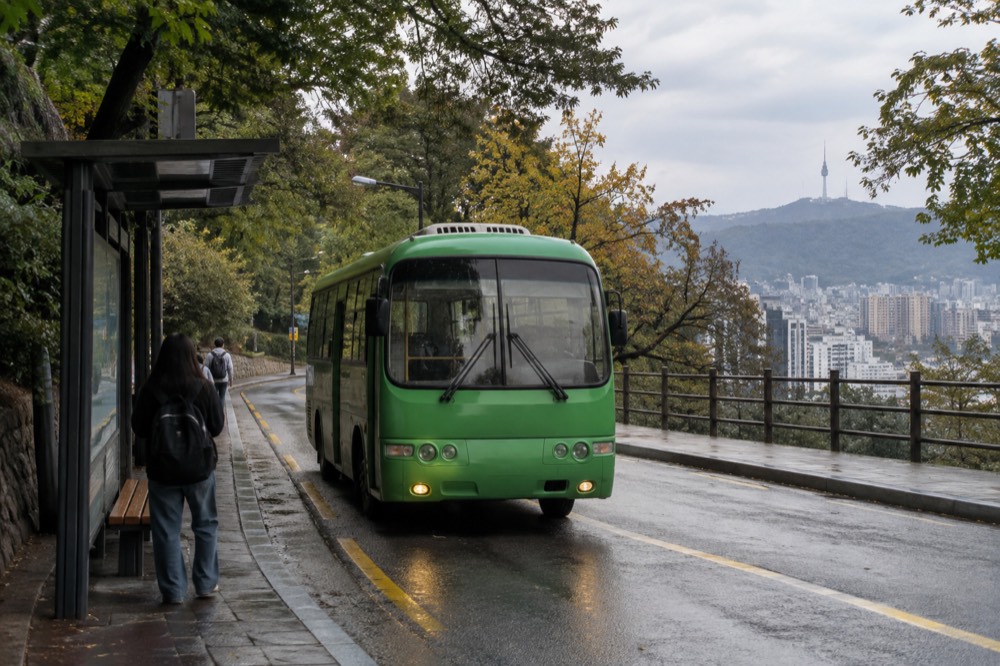 A small green shuttle bus at a stop on a wooded mountain road above city buildings