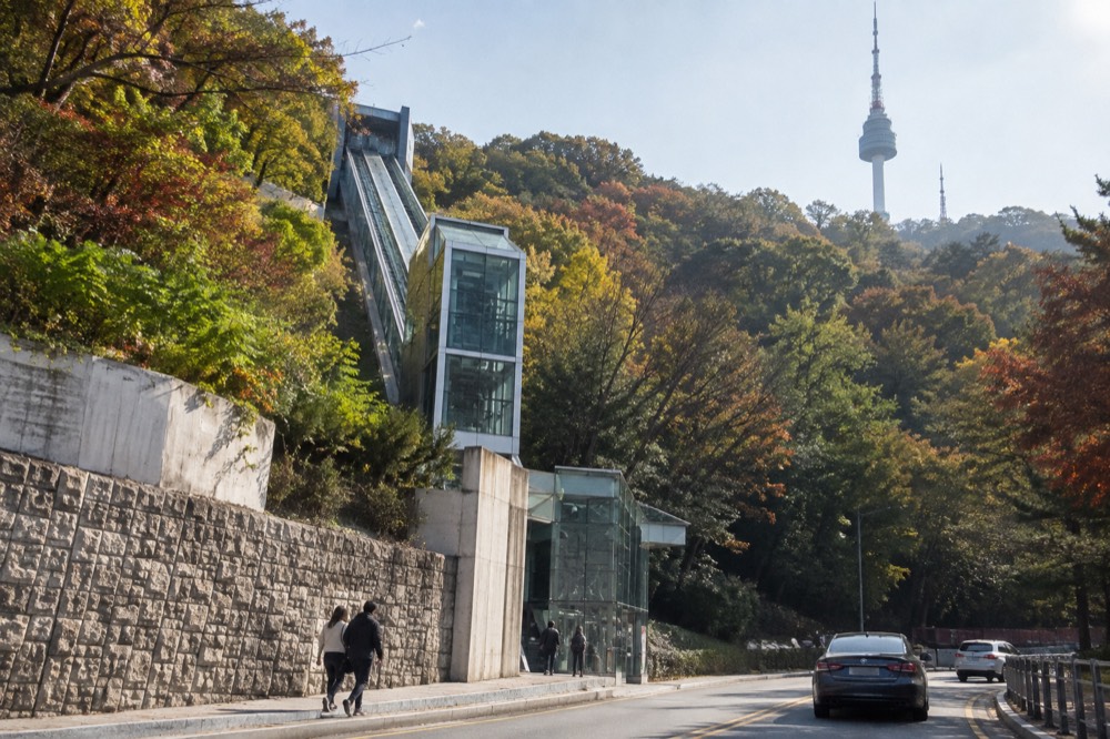 Glass-walled inclined elevator structure rising along the Namsan hillside beside the road