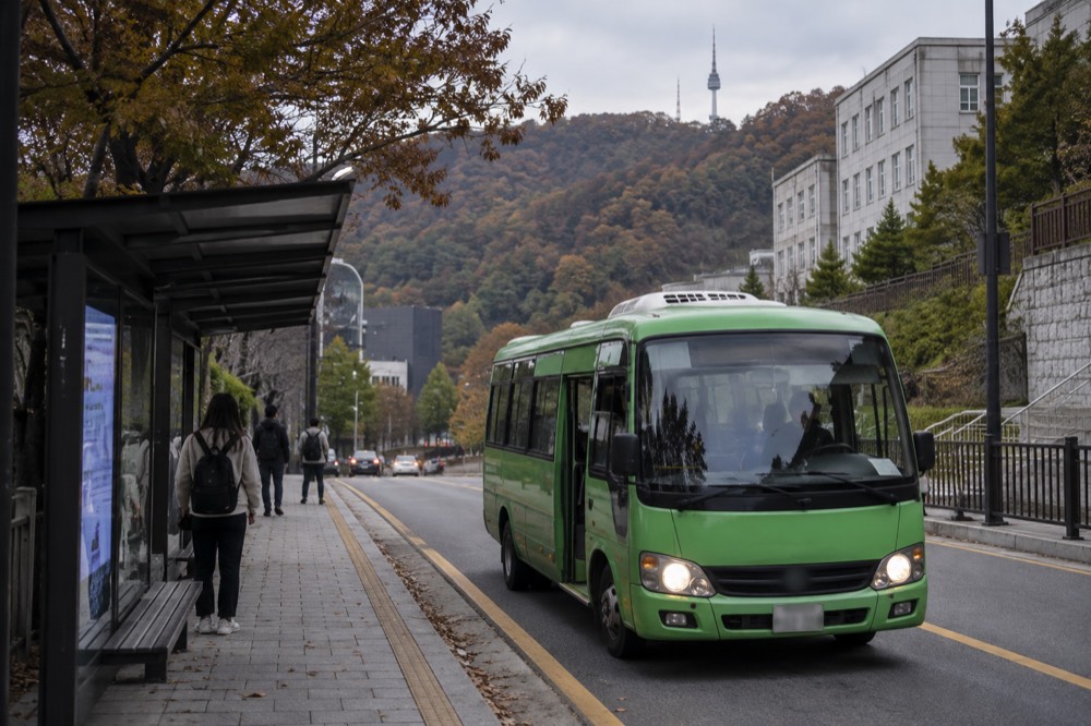 A small green shuttle bus parked on a narrow road with wooded slopes behind it