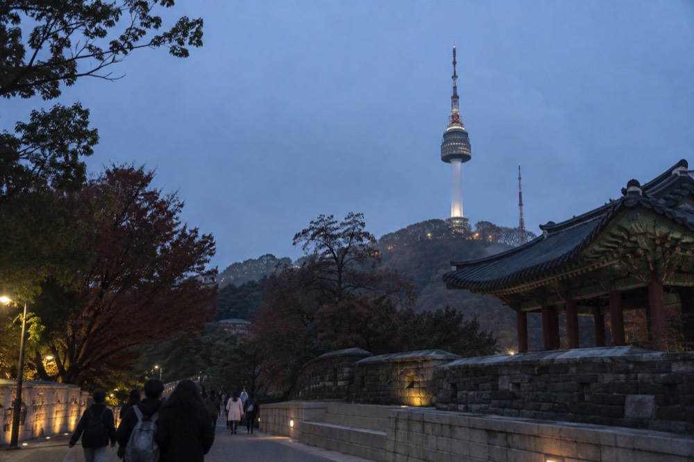 A tall tower with a lit observation pod standing above a tree-covered summit at dusk