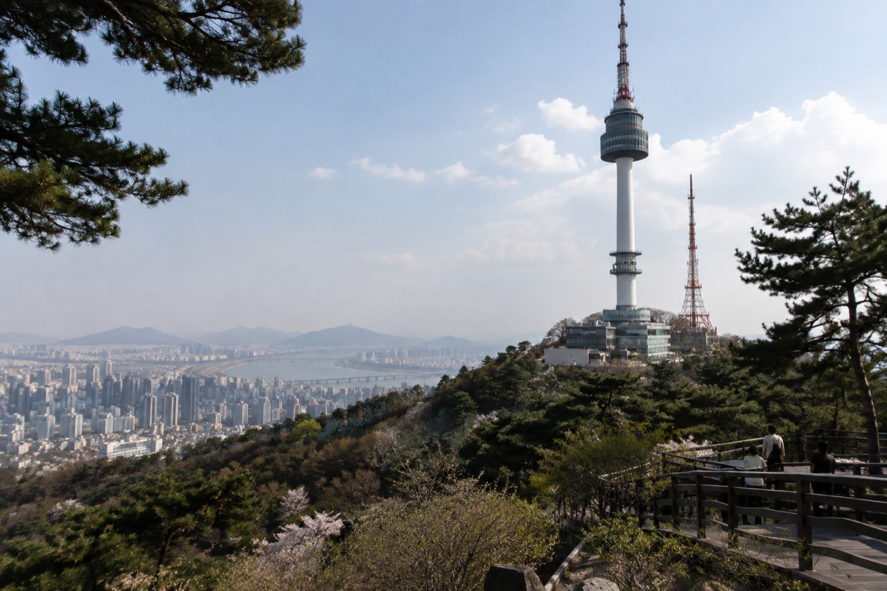 N Seoul Tower rising above the tree-covered Namsan summit under a wide sky, with the Seoul skyline spread out behind it
