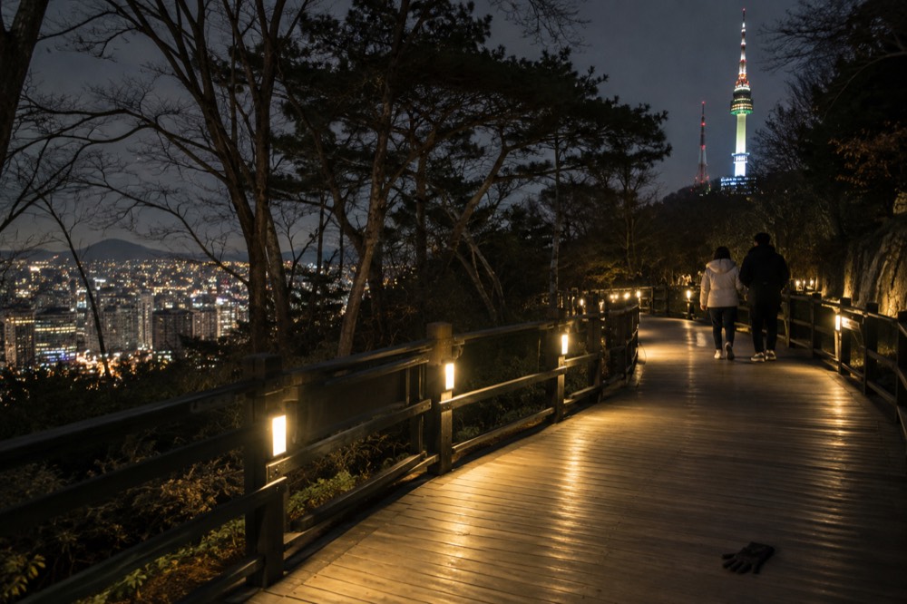 A lit timber deck trail on Namsan winding through trees with the city glowing below
