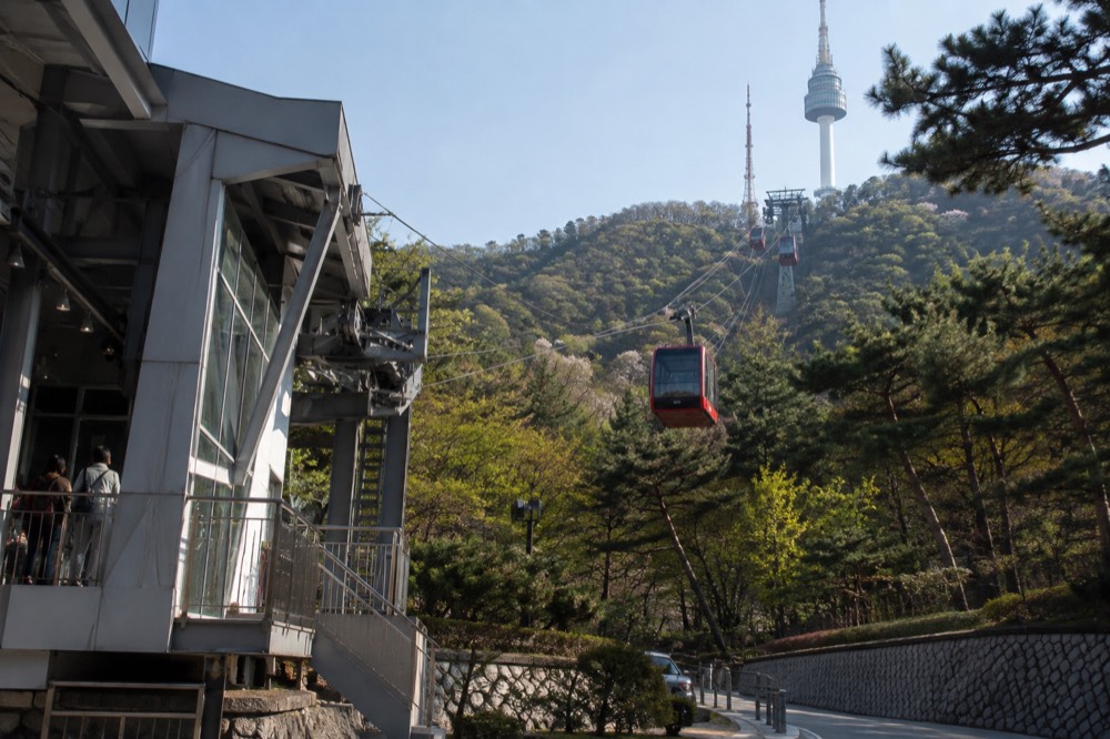 Cable car cabins travelling up a forested hillside toward a tower on the summit above city buildings