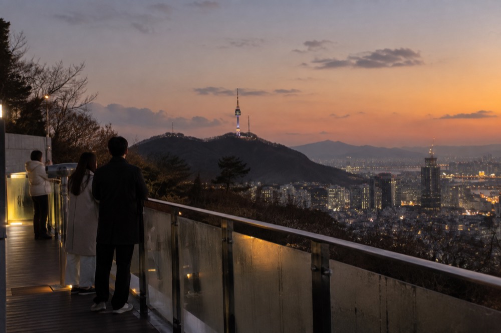 A city skyline at dusk seen from a hilltop terrace with railing in the foreground