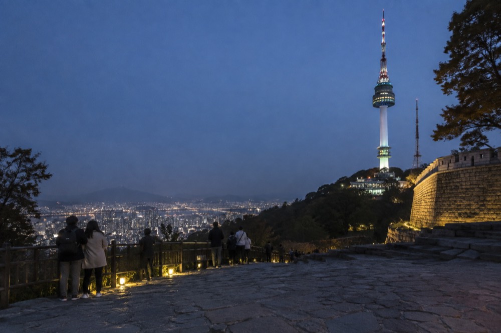 A lit communications tower on a forested hill above evening city lights