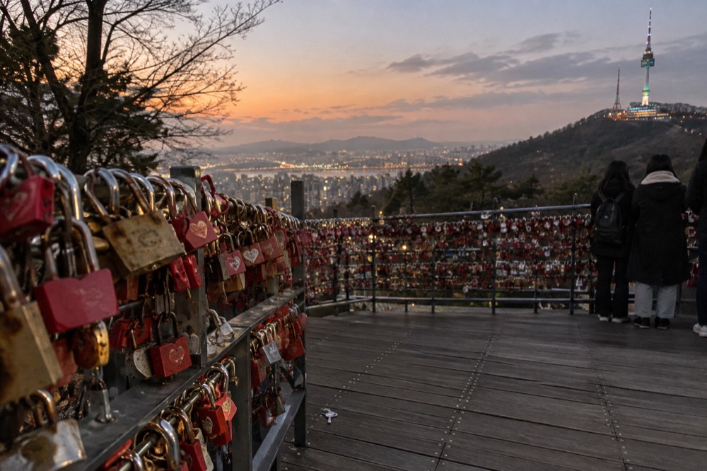 Rows of padlocks attached to metal railings on an open terrace overlooking a city