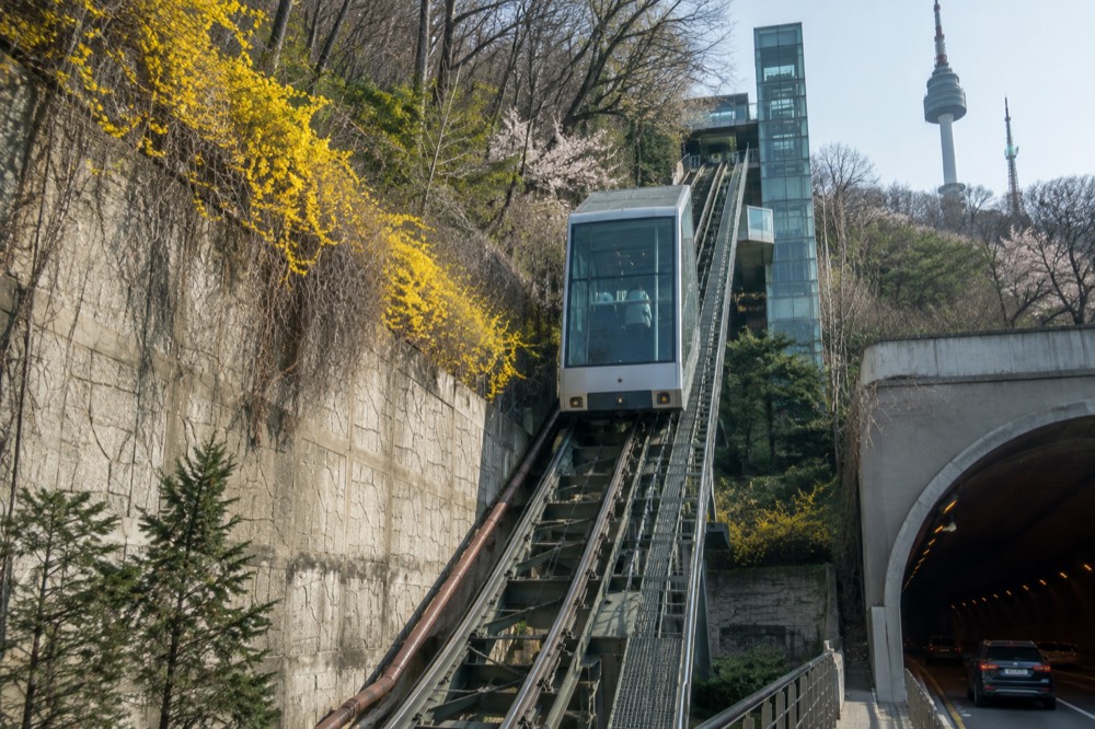 Glass-walled inclined elevator ascending a steep planted slope beside a Namsan tunnel entrance