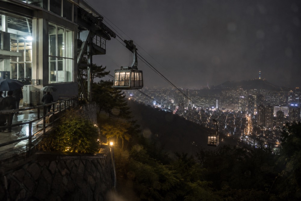 Cable car cabins moving along the lit ropeway line above the Myeongdong side of Namsan at night