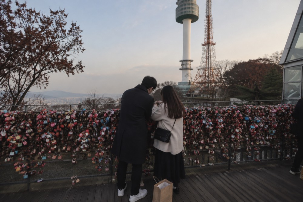 A couple attaching a small padlock to a railing covered in existing locks, with the tower structure rising behind