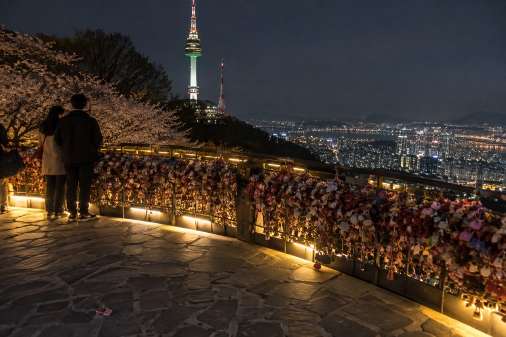 A hilltop terrace at night with illuminated railings and a glowing tower above city lights