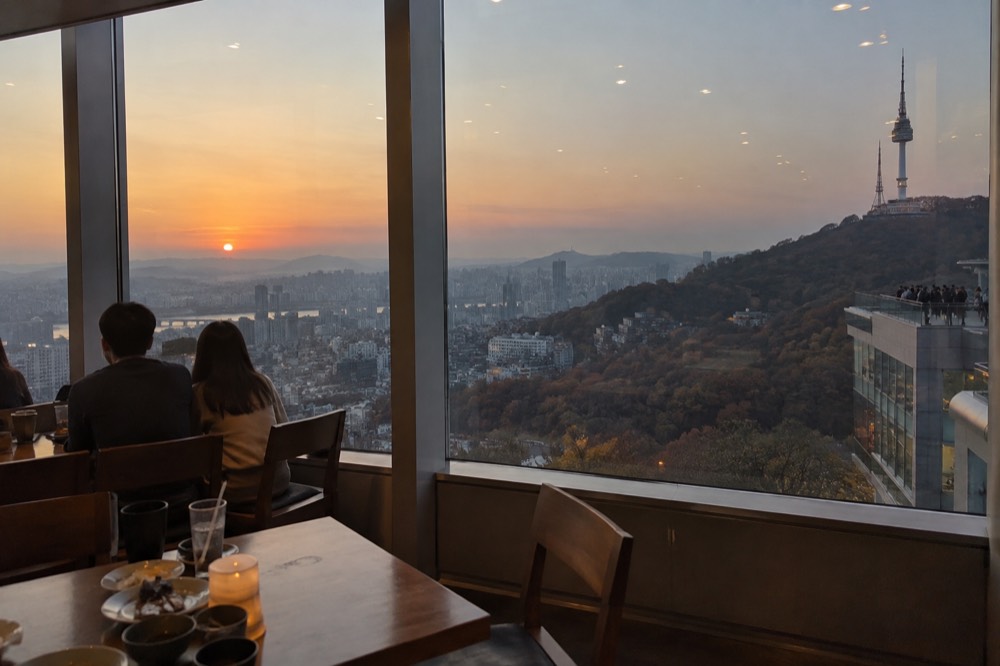 Dining tables set beside a large window with the Seoul cityscape at dusk