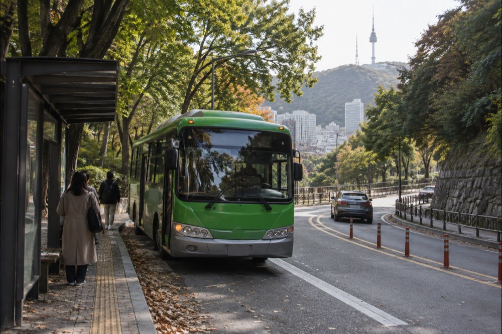 Green shuttle bus at a stop on the Namsan circular road with forest on both sides