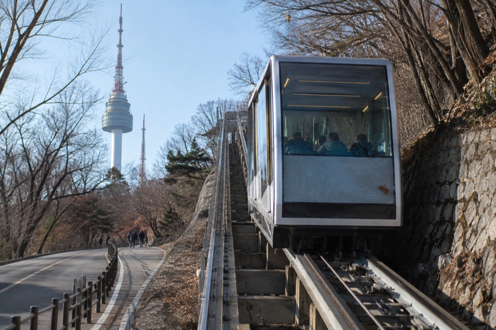 Glass-walled inclined elevator rising along the slope beside the Namsan roadway