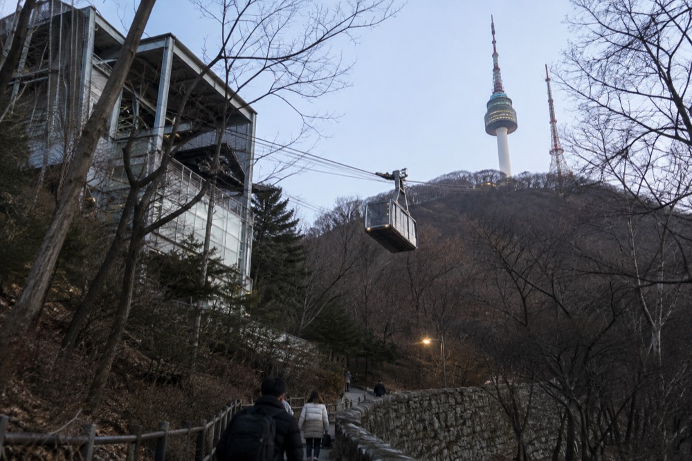 A cable car cabin ascending a steep forested slope toward a tower on the ridge above