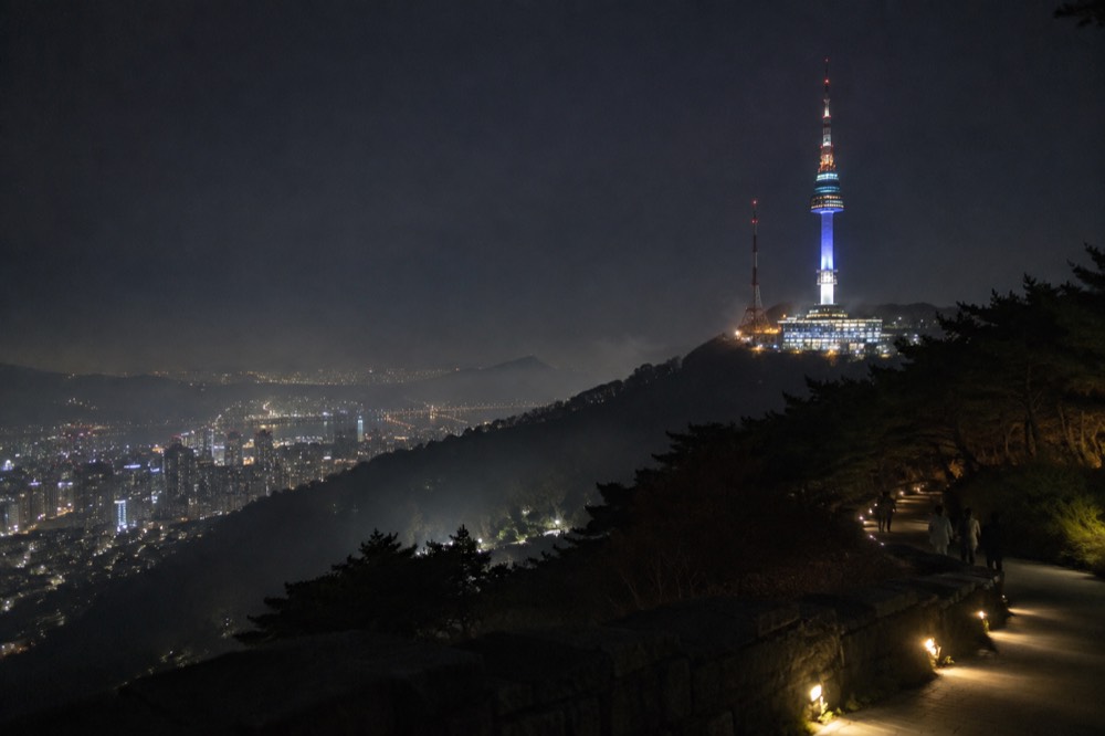 Illuminated tower standing above dark forest with city lights spread across the valley below