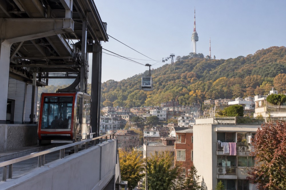 Cable car cabins suspended on a line rising above dense apartment rooftops toward a forested hilltop