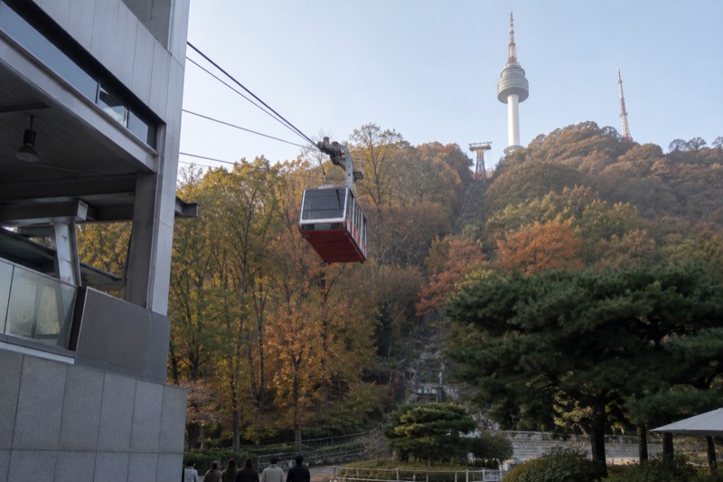 A cable car cabin suspended on its line, climbing a forested slope toward a tower on the ridge.